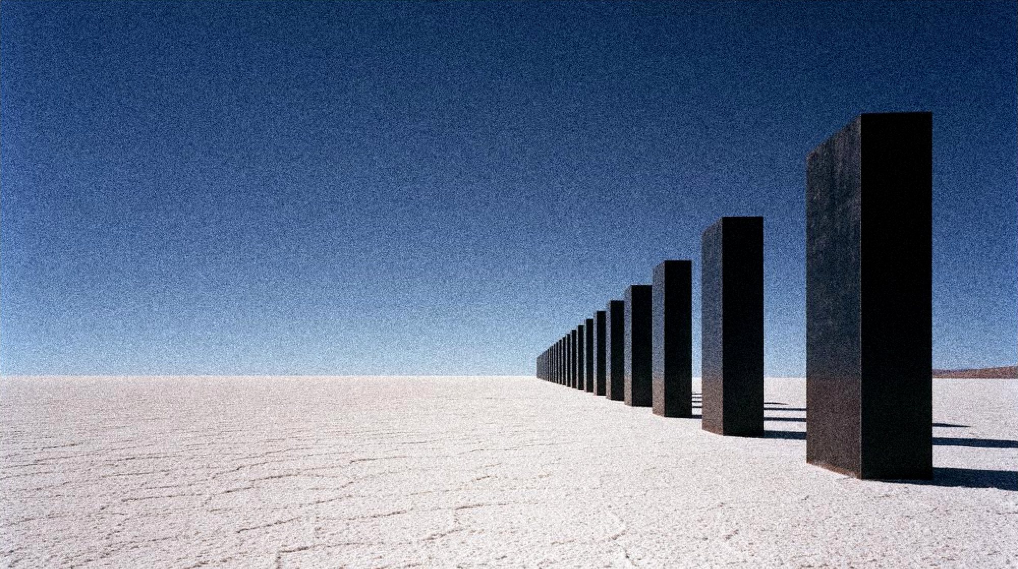A row of black slabs receding across a white salt flat to the horizon